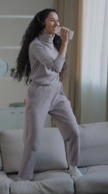 Evdeki rahat koltukta dans eden çekici, genç ve komik bir kızın dikey görüntüsü. En sevilen yüksek sesli müzik dinamik hareketleri. Şarkı söylemek. Cep telefonunu mikrofon olarak tut.