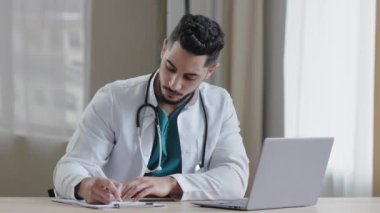 Yoğunlaşmış Arap erkek doktor pratisyen hekimi klinik kabinesinde oturup medikal sosyal uygulamalarda uzak sohbetler yapar. Online danışmanlık yapar, reçete yazar, reçeteli arama bilgileri yazar.