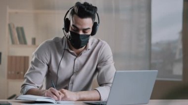 Tıp maskeli genç Arap erkek yazar kulaklık takıyor. Salgın sırasında evde oturup kitap çalışmalarında online eğitim Webinar 'ı günlük ev ödevi yazarken bilgisayar kullanıyor.