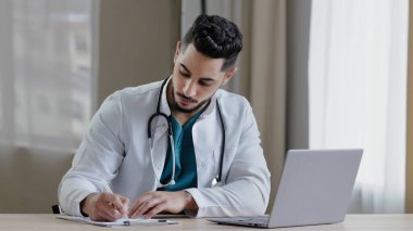 Yoğunlaşmış Arap erkek doktor pratisyen hekimi klinik kabinesinde oturup medikal sosyal uygulamalarda uzak sohbetler yapar. Online danışmanlık yapar, reçete yazar, reçeteli arama bilgileri yazar.