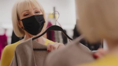 Giysi mağazasında koruyucu maske takmış olgun bir kadın aynaya bakar ve şık bir elbise dener. Kadın alıcı yeni kıyafetler seçer. İndirimli kıyafet satışı için ürünler seçer.