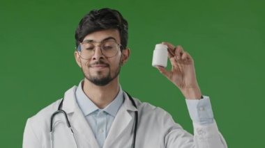 Yeşil arkaplan üzerinde duran Hintli erkek doktor danışmanı kamera şişesinde ağrı kesici haplar vitamin hapları ve alerji tedavisi için ilaç öneriyor.