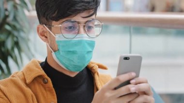 Yakın plan koruyucu maskeli genç Arap adam akıllı telefondan mesaj yazıyor mobil uygulama kullanıyor sohbet sosyal ağları kullanıyor mesaj yazıyor telefon tarayıcı ağından online sipariş veriyor modern teknolojiden keyif alıyor.