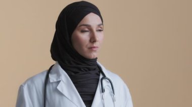 Üzüntülü müslüman genç kadın doktorun portresi İslami jinekolog başörtüsü takan doktor düşünceli kadın eczacı rüya gören kız bej arka planda duruyor.