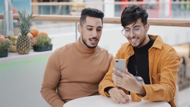 カフェに座っている2人の幸せなアラブの友人ブロガースマートフォン画面を見てウェブカメラを介して通信する携帯電話でビデオブログを記録するモバイルアプリケーションを使用してソーシャルメディア上でビデオを作る自撮り — ストック動画