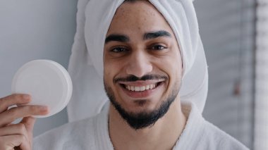 Kafasında krem kavanozu olan Arap İspanyol asıllı adam kameraya bakıp gülüyor banyoda doğal erkek kozmetik ürünleri öneriyor yüz bakımı nemlendirici tedavisini öneriyor.
