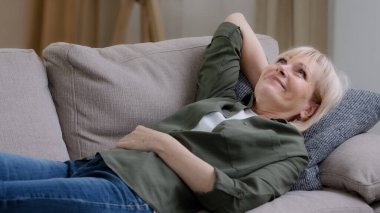 Mutlu, orta yaşlı, düşünceli, düşünceli bir kadın evdeki kanepede dinleniyor. Huzurlu bir ruh hali, uykulu, tembel, yaşlı, olgun bir kadın rüyası. Oturma odasında rahat bir kanepede uzanıyor. Stres yok.