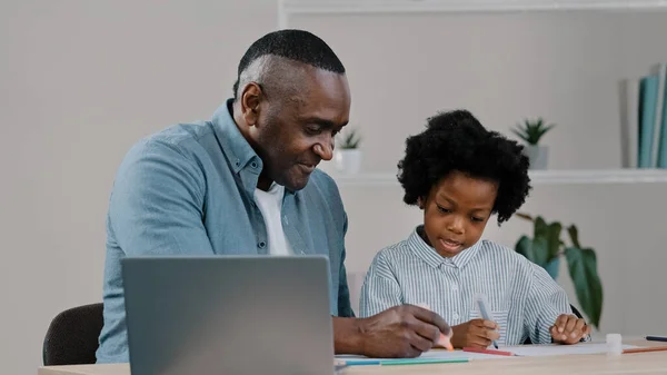 Adult father home teacher helping little daughter to do homework sit at desk in room study remote using laptop man explaining kid girl task african american family having fun together laughing smiling