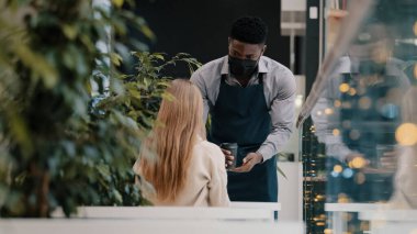 Önlüklü, tıbbi maskeli, genç, profesyonel bir garson. Kafede müşteriye kahve ikram ediyor. Kâğıt bardakta kahve getiriyor.