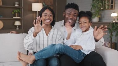 Kızı olan Afrikalı bir aile evde oturmuş el sallıyor kameraya bakıyor ve kameraya bakıp merhaba diyor. Web kamerası görüntüsü Amerikalı ebeveynler ve çocuk videosu çevrimiçi video görüşmesi vlog sohbeti