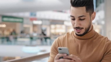 Yakın plan genç Arap adam alışveriş merkezinde elinde telefonla dolaşıyor internetteki arkadaşlarıyla sohbet ediyor e-posta cevapları okuyor çevrimiçi mağazalarda sipariş veriyor sosyal ağda ücretsiz kablosuz iletişim kullanıyor.