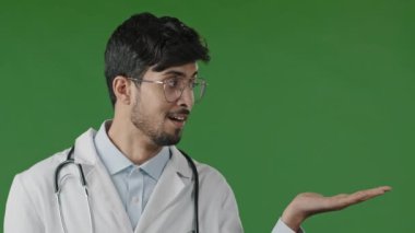 Arap Hindistanlı genç doktor pratisyen adam, reklam için iş yeri tanıtım yeri olan palmiye üzerinde fotokopi alanı tutuyor. Yeşil arkaplan üzerinde izole edilmiş hayali bir nesne duruyor.