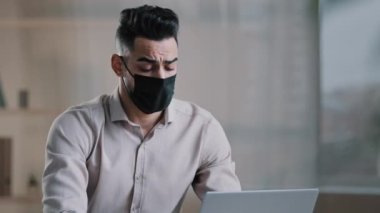 Hasta İspanyol asıllı, bunalmış genç erkek, tıbbi maskeli iş adamı yorgun, yorgun, motivasyonsuz, sıkıcı Arap adam, salgın sırasında bilgisayar başında çalıştıktan sonra baş ağrısı çekiyor.