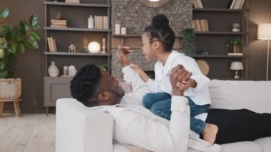 Afrikalı baba evde kanepede yatıp kızıyla oyun oynuyor. Birlikte boş vakit geçiriyor. Üzgün küçük kız çocuğu eğlendiriyor.