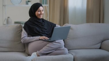 Dost canlısı Arap müslüman kadın tesettür koltuğunda oturup sohbet videosu izleyerek bilgisayar kamerasından arkadaşlarınızı selamlayın sosyal ağlarda sanal iletişim kurmak için evdeki wi-fi kullanın.