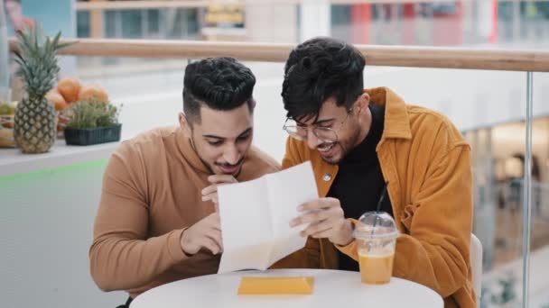 カフェに座っている2人の若いインド人学生は紙の手紙を手に入れますショックを受けた幸せな男性良いニュース通知を読む喜びの友人は勝利を祝う喜び優れた結果友好的なサポート — ストック動画