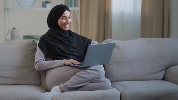 フレンドリーなアラビア語イスラム教徒の女性の女の子でHihab座っ居心地の良いソファトークビデオ通話で友人挨拶でコンピュータウェブカメラ取るインタビュー距離仮想通信のソーシャルネットワーク使用家でWi — ストック動画