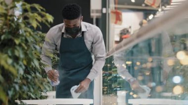 Önlüklü genç erkek garson, dezenfektan spreyleri temizliyor. Kafe restoranını, tıbbi maske temizleme kafeteryasında yarı zamanlı çalışan ciddi bir Afro-Amerikan öğrencinin açılışına hazırlıyor.