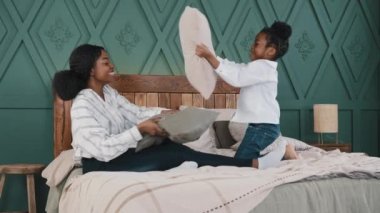 Mutlu Afrikalı anne, bebek bakıcısı ablası, tatlı kız çocuğu, yatakta oturuyor yastık savaşı yapıyor, evde eğleniyor, anne ve çocuk, komik dövüş oyunları oynuyor, sabah kahkahası atıyor.