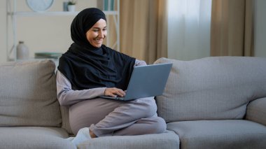 Dost canlısı Arap müslüman kadın tesettür koltuğunda oturup sohbet videosu izleyerek bilgisayar kamerasından arkadaşlarınızı selamlayın sosyal ağlarda sanal iletişim kurmak için evdeki wi-fi kullanın.