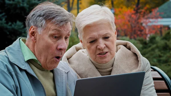 Shocked upset elderly married couple partners looking at laptop screen sit in autumn park disappointed old woman read bad news worried mature family feel stress discuss problem grandfather cover head
