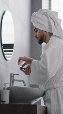 Kafasında havluyla banyo cüppesi olan yakışıklı Arap sakallı adam sıvı cilt bakımı kullanıyor. Yüzü tonik suyla temizliyor. Aynaya bakıyor.