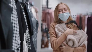 Medikal maskeli mutlu müşteri bayan müşteri alışveriş merkezinde beyaz kadın alışveriş meraklısı kadın kıyafetlerle dans ederken Kara Cuma günü indirimli indirimler yapıyor.