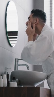 Dikey görüş Arap İspanyol, bornozlu sakallı adam banyodaki aynadaki yansımaya bakar yüz derisini krem nemlendirici kozmetik ürünler anti-akne doğal cilt bakımı