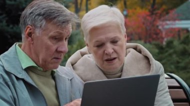 Beyaz tenli yaşlı çift, olgun bir eş, açık hava bankında oturuyor, yaşlı eşler dizüstü bilgisayarda wi-fi kullanıyor, bilgisayar ekranı alışverişine bakıyor, internet üzerinden bilgi topluyor, internet üzerinden sipariş veriyor.