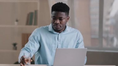 Afrikalı meşgul erkek işçi Amerikan işadamı öğrenci ofiste oturup bilgisayarla yazma işini bitiriyor günlük işleri yapıyor bir bardak soğuk su içmek sağlık dengesini koruyor.