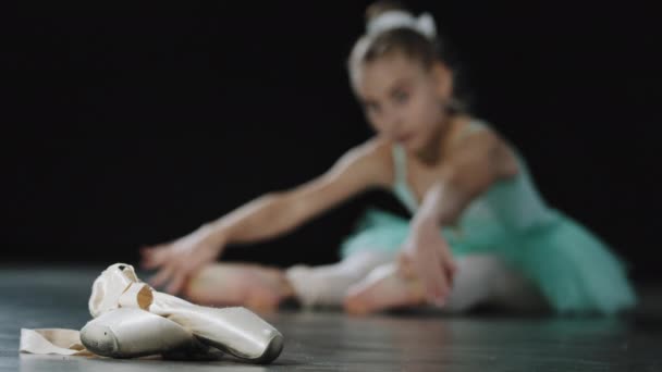 ブルーチュチュの10代のバレリーナの女の子が床のストレッチに座って バレエシューズは前景に靴を指します 稽古後に休んだ子どもたちがストレッチの足を学び 舞台に立つ準備をする — ストック動画