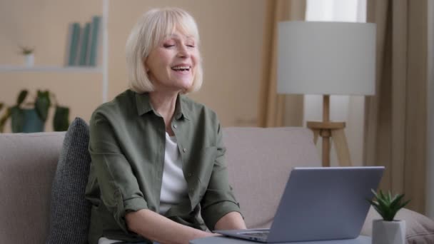 白人成熟した老婦人は自宅でソファの上に座っているノートパソコンのカメラに手を振っこんにちはビデオ通話会議チャットで孫とのリモート会話でウェブカメラのコンピュータアプリケーションオンライン — ストック動画