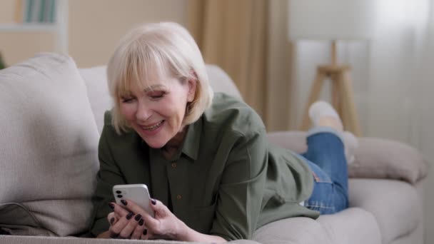 古い幸せな白人の祖母60歳年上の女性50代の女性ブロンド年上の女性は自宅でソファに横たわっていますリモートモバイル会話ビデオ通話会議チャット笑顔電話カメラと話して — ストック動画