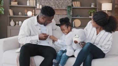 Çift ırklı aile oturma odasında kanepede oturuyor ve hafta sonunu birlikte geçiriyorlar. Afrikalı anne ve baba ve kız çocuğu evde eğleniyorlar. Baba küçük kızı kanepede gıdıklıyor. Gıdıklıyor. Oyun oynuyor.