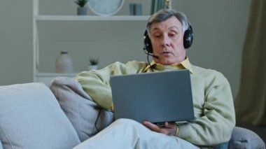 Evdeki kanepede oturan 60 'lı yaşlardaki erkek dijital kulaklık takıyor ve konferans için bilgisayar uygulaması kullanan yaşlı bir dizüstü bilgisayarla konuşuyor. Kulaklık takmış yaşlı büyükbaba uzaktan konuşuyor.