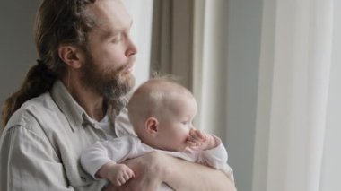 Yakın plan beyaz baba sakallı bebek kızını kucağında tutuyor yeni doğmuş bebeği pencereden dışarı bakıyor babası evde oynayan kısa sarı saçlı çocuğa üflüyor. Babalık koruması