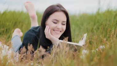 Genç, dingin, beyaz kadın çimlerin üzerinde uzanan çekici, gülümseyen ergen kız kitabı okuyor. Romantik edebiyat tarihinin tadını çıkarın. Dışarıda rahatlayın.