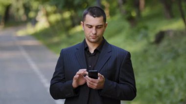 Ciddi yetişkin beyaz adam parkın dışında duruyor. Siyah resmi kıyafetler giyiyor. Cep telefonu kaydırma sayfalarını akıllı telefon ekranında tutuyor.