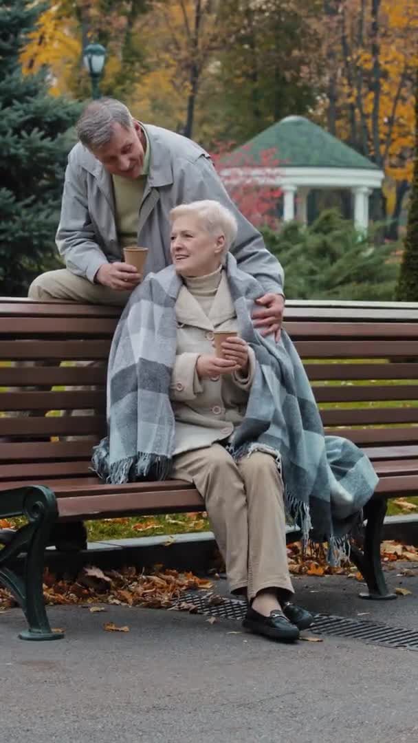 幸せなカップルの垂直ビュー白人家族のシニア男性ベンチに女性の後ろに座る巨大な高齢者の成熟した妻は秋の公園の外で一緒に快適に感じる熱いコーヒーを飲む楽しい会話を楽しむ — ストック動画