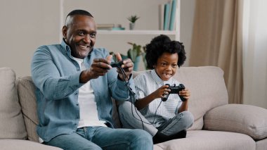 Mutlu Afro-Amerikan aile babası ve kızı birlikte iyi eğlenceler. Babası, kız çocuğuyla konsolda video oyunu oynuyor. Kontrolör, kanepeye oturup yarışmayı kazanırken eğleniyor.
