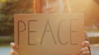 Kapalı kadın elleri kartondan yazılı barış sembolü, çekici genç kadın, güzel kızıl saçlı kız parkta duruyor ve sloganı protesto eden barışçıl afiş anti-mağdur pankartını gösteriyor.
