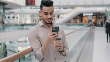 Arap adam, tişörtlü, kahve içen Hintli işadamı cep telefonu ekranına bakıyor, akıllı telefonuyla sohbet ediyor, ofis içi alışveriş merkezinde kağıt bardaktan kahve içiyor.
