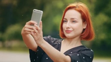 Portre stili güzel bir kız beyaz saçlı çekici kızıl saçlı kadın blogger model parkta duruyor akıllı telefon cep telefonuyla fotoğraf çekiyor mobil kamera kayıtları video blogu için video çekiyor.