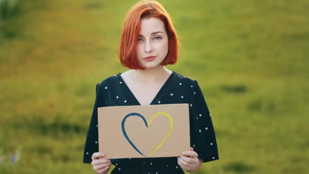 悲伤的红头发女人拿着彩绘的纸板画蓝色的黄色爱情的心形浪漫的象征表现柔情迷人的年轻漂亮的姑娘站在室外阳光下 — 图库视频影像