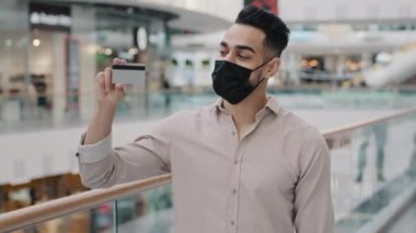 Arabian Indian Hispanic man in medical mask standing with purchases. Arab millennial guy customer holding credit bank card and shopping bag enjoys sale happy with discount male buyer posing indoors
