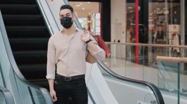 Arap İspanyol erkek tüketici müşteri müşteri müşteri işadamı tıbbi maske takan alışveriş torbaları takan alışveriş merkezindeki yürüyen merdivenlerde duran indirimli satış sonrası poz veren
