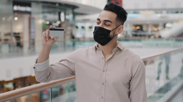 医療用マスクに身を包んだアラビア系インド人ヒスパニック男性 アラブ千年男顧客保持クレジットカード銀行カードとショッピングバッグ楽しんで販売幸せな割引男性バイヤーポーズ屋内 — ストック動画