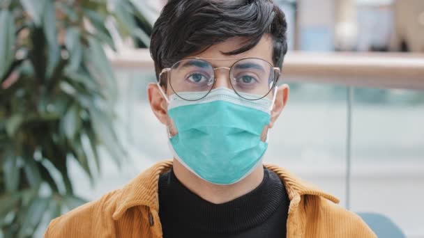 屋内に立つ保護医療用マスクの肖像クローズアップ若いインド人男性カメラの個人的な安全性を見てポーズパンデミックが発生したため 感染症は意識的に健康サイン意識を保護します — ストック動画