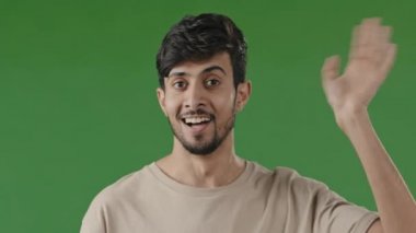 Indian cheerful man say hi greeting waves male hand friendly arab hispanic guy handsome boyfriend demonstrates welcome hello goodbye sign shakes wrist palm smiling stands on green chromakey background