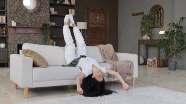Young crazy funny carefree woman lying upside down on cozy sofa in living room shaking arms and legs dances lying enjoys being alone at home on weekend celebrating victory resting relaxing leisure